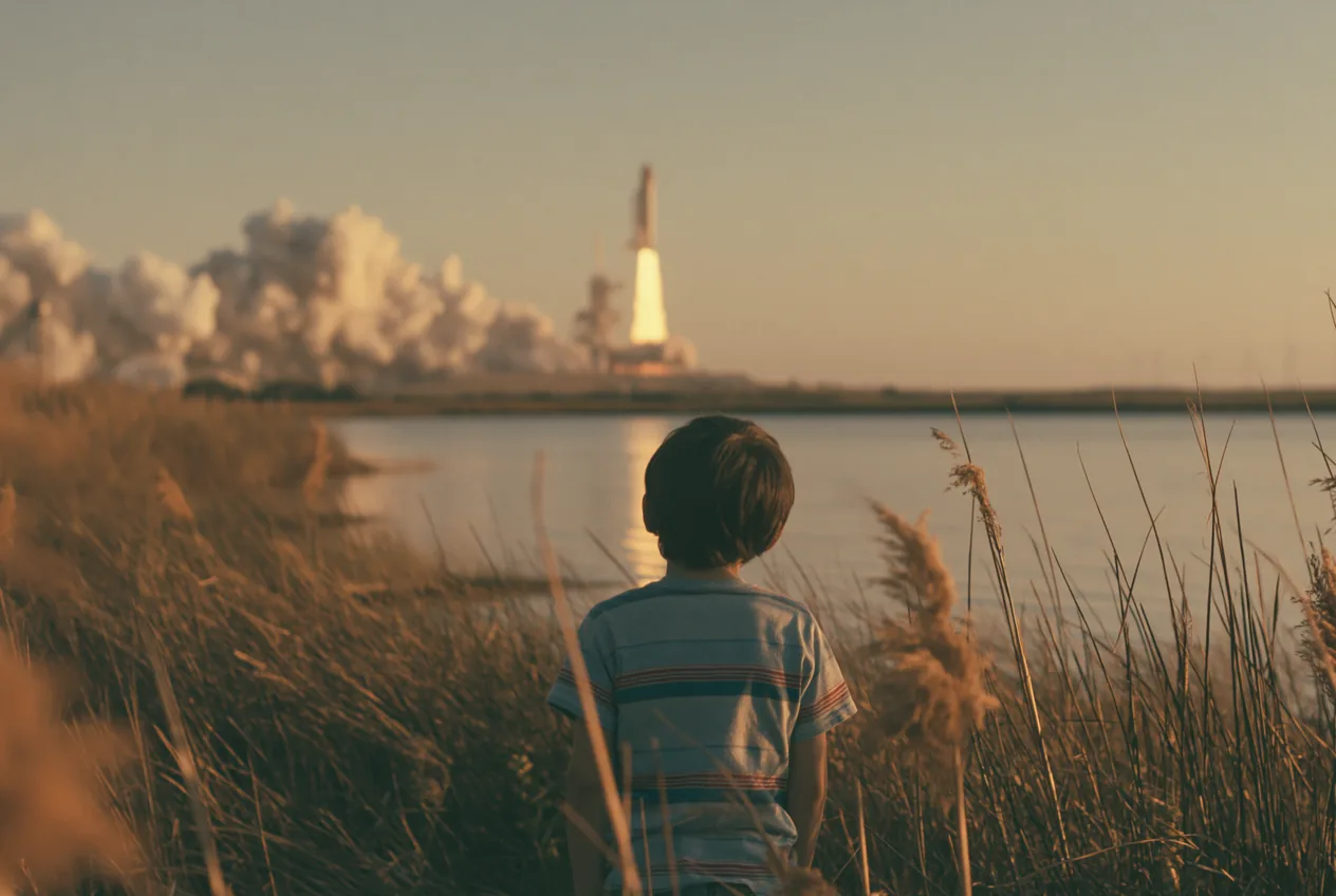 Un enfant de dos, assis dans les hautes herbes au bord de l’eau, regarde une fusée décoller à l’horizon dans la lumière du soir.